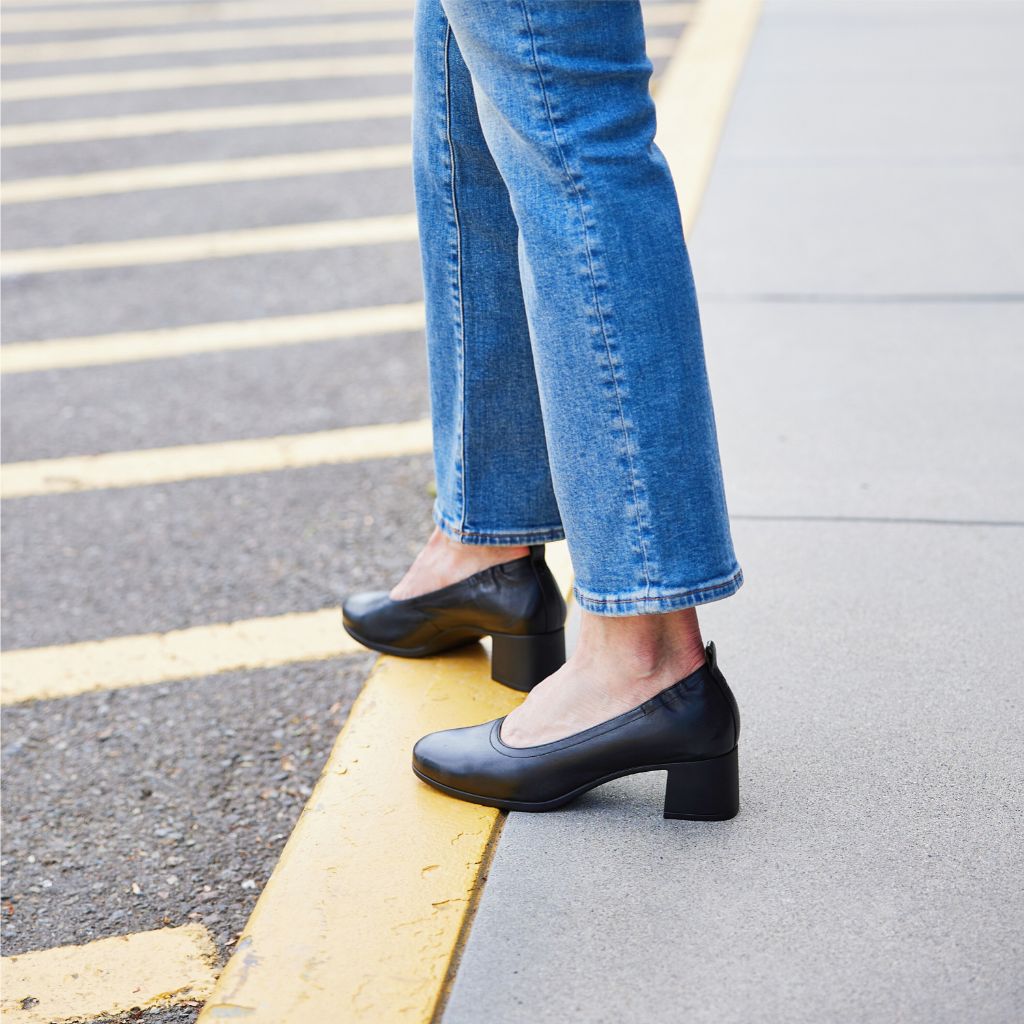 Woman wearing black PIAR Performance Heels standing on yellow curb in jeans