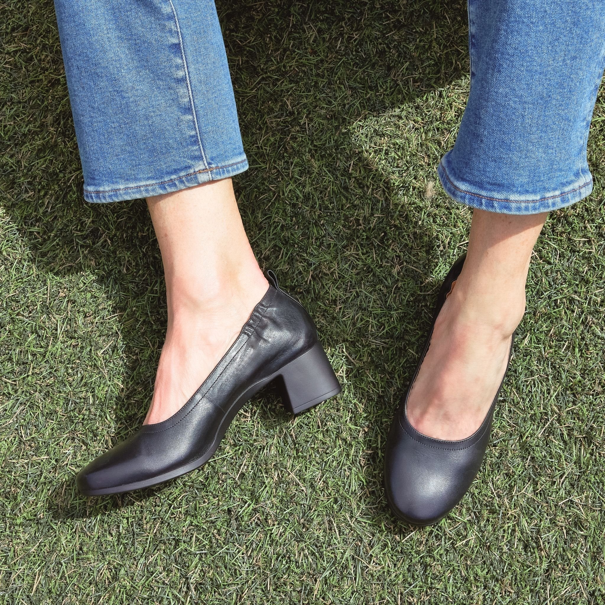 The Performance Heel by PIAR - Woman Wearing Stylish and Comfortable Black Leather Block Heel Pumps Sitting on Grass